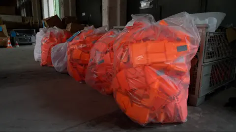 BBC Large numbers of bright orange lifejackets are seen in plastic bags inside a warehouse