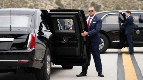 Getty Images Two US Secret Service agents, both in suits, one in sunglasses, hold open the back passenger doors on two versions of the "Beast" armoured limousine.