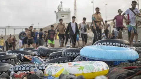 Anadolu via Getty Images Rubber flotation rings sit on a beach in front of a line of arriving migrants in Ceuta.