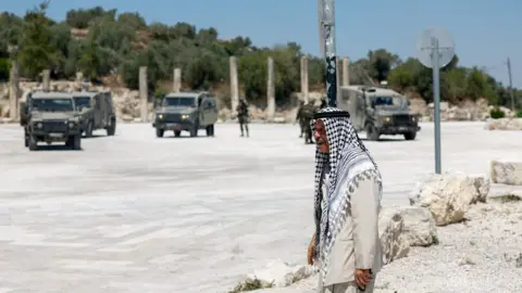 Reuters A Palestinian man walks near Israeli troops at the archaeological site of Sebastia, in the occupied West Bank (29 June 2026)