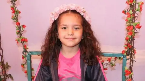 Family/Reuters Hind Rajab smiles at the camera. She has long, brown wavy hair and is wearing a pink flower crown on her head. She also has a pink T-shirt and black zip-up on. She is sitting on a chair adorned with garlands of red flowers.