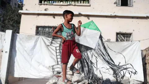 NurPhoto via Getty Images A Palestinian boy holds a kite at a displacement camp in Gaza City (23 August 2026)