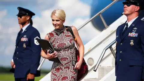 Getty Images Natalie Harp walks off Air Force One wearing a sleeveless printed red dress. She is smiling and has a laptop and several phones in her hands.