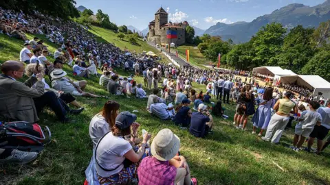 AFP via Getty Images Hundreds of people gather on the slopes below Vaduz Castle, the palace and official residence of the Prince of Liechtenstein, during its National Day celebrations in Vaduz, on 15 August. Mountains can be seen in the background.