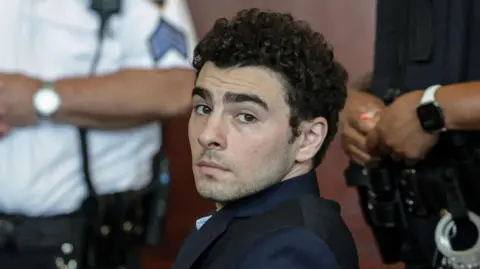 Getty Images Luigi Mangione, dressed in a dark suit and blue chequered shirt, looks at the camera with law enforcement officials in the background