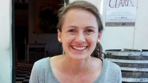Facebook A woman with long brown hair, tied back in a ponytail, smiling at the camera. She is wearing pearl-like earrings and a scoop neck grey top.