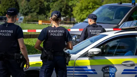 EPA Three police officers in uniform, including a bulletproof vest reading Kantons Polizei at the back, stand watching a field cordoned off by white and red tape. A fourth officer sits inside a police car parked next to his colleagues