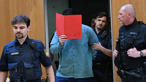 Reuters Farhad N holds a red folder over his face as he is brought into court by police officers in Munich on 6 August 2026.