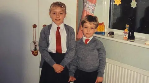 Dalibor Sentic The childhood photo of Claire and Peter which was sent to Dalibor Sentic in the care package he recieved in 1992. Claire and Peter are photographed in school uniforms, stood in what appears to be a bedroom. Claire is taller than Peter and stood to the left of the image. She is wearing a black skirt, white blouse, red tie, and grey cardigan. She has blonde hair with a fringe. Her hair is tied up. Peter is shorter than Claire and is photographed to the right of her. He is wearing black trousers, a white shirt and red tie, with a grey jumper over the top.