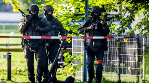 EPA Three police officers in riot gear (helmet, bulletproof vest, knee and elbow protection) carrying guns patrol on the street, a police cordon is visible in the foreground