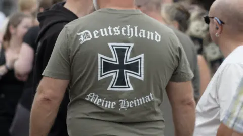 BBC The black of a man wearing a khaki green t-shirt which says 'Deutschland, Meine Heimat' written in a Gothic-style font. The t-shirt shows the insignia of the iron cross 