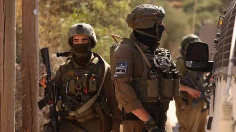 EPA Two IDF soldiers in military gear and face coverings block a road near the West Bank village of Qusra near Nablus. One soldier is seen facing towards the side of a van while a second holding a rifle is facing towards the camera but looking off in the distance.
