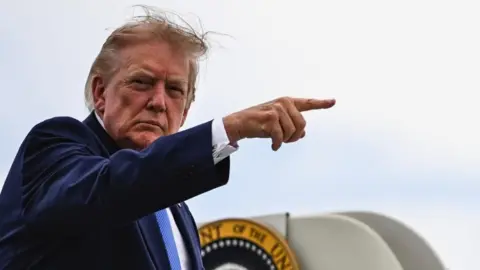 Reuters Donald Trump lifts his right arm up and points with his index finger. Part of the presidential seal can be seen behind him. He is looking to his right at the camera.