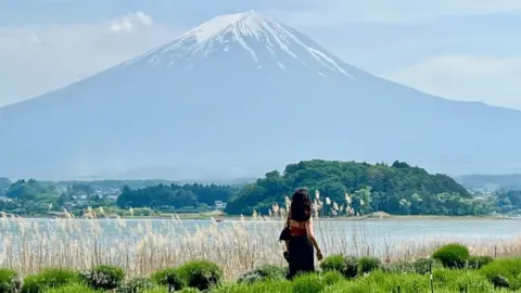 Maitri Sanghvi A photo taken from behind of a woman looking up at a snow-topped Mount Fuji, with a lake in front of it