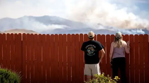 Getty Images Two people look over what appears to be a domestic fence. They are looking at where smoke is rising from hills in the background. 
