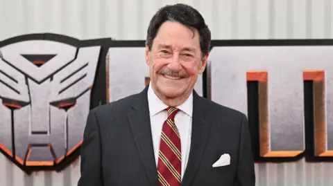 Getty Images Peter Cullen standing and smiling in front of a Transformers logo backdrop