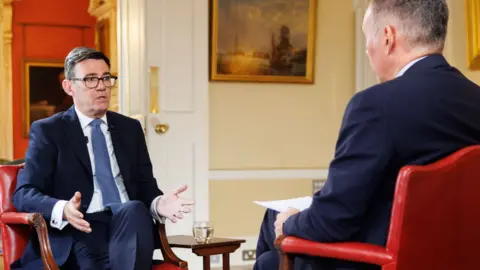 Simon Dawson / 10 Downing Steet Andy Burnham wears a dark suit sitting on a chair speaking with Roger Johnson who wears a dark suit. They are both sitting on red chairs.