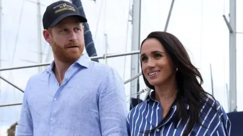 Reuters Harry and Meghan seen walking together in Sydney harbour in April. He is wearing a blue shirt and a cap, she is wearing a striped blue shirt.