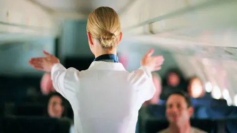 Getty Images An air stewardess facing away from the camera shows passengers on a jet the emergency exist during a safety briefing. She is wearing a white shirt and her blonde hair is in a tight bun