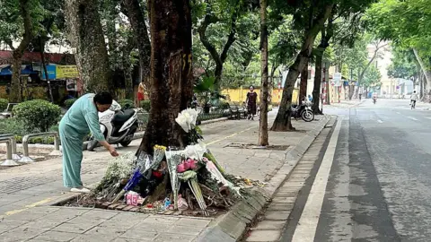 Getty Images A woman in mint green clothes bends over a ring of flower bouquets arranged around the trunk of a roadside tree
