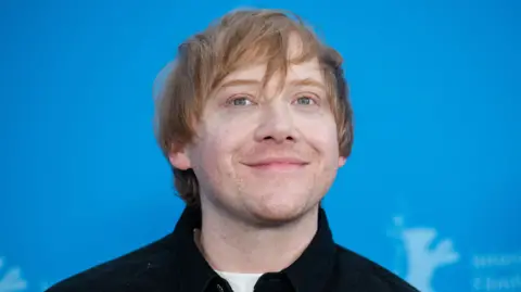 Getty Images Rupert Grint in a dark shirt and white T-shirt, smiling and posing in front of a blue background