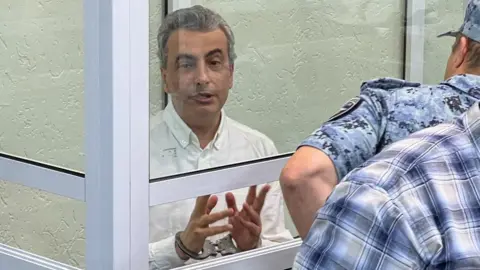 Reuters Lev Shlosberg (left, in handcuffs) attends a court hearing in Pskov, western Russia. Photo: 17 September 2026.