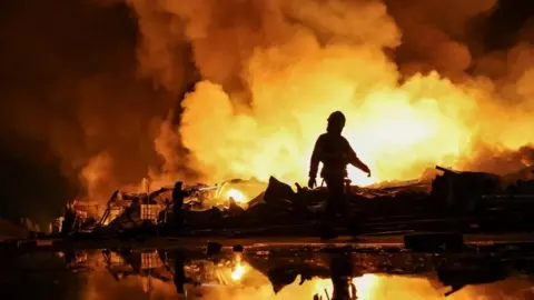 Reuters Firefighters work at a site of a Russian missile strike, their silhouettes contrasted against the glowing yellow and orange of smoke plumes illuminated by large fires. 
