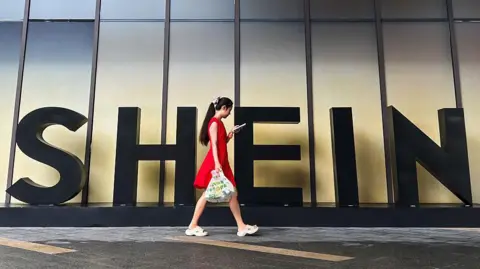 Getty Images A woman in a red dress looks at her smartphone while walking past a Shein sign outside the company's office in Guangzhou, China. 
