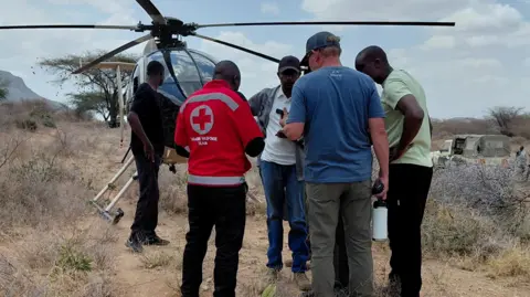 Kenya Red Cross/Handout via REUTERS Five men stand on dusty arid dirt with a helicopter in the background. One of them has a Red Cross top on.