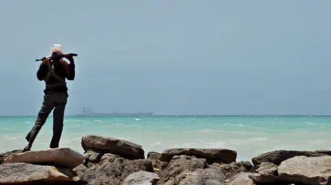 AFP via Getty Images A man - with his back to the camera - stands on some rocks to the left of the picture. Holding a gun across his shoulders, he is looking out to sea where a tanker is in the far distance. 