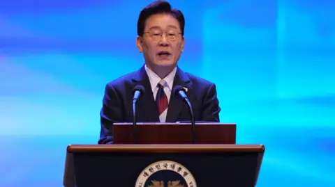 Getty Images Lee Jae Myung speaks at a lectern during a Liberation Day ceremony. He wears a dark suit and glasses, in front of a blue backdrop.