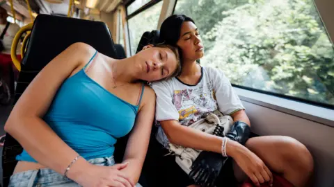 Getty Images Two young women asleep on a train - one wearing a blue vest top leans on the other's shoulder