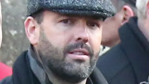 Collins Photo Agency Daniel Kinahan, a man with short dark hair and a stubbly beard, pictured at a funeral in 2016.  He is wearing a grey peaked cap, a black shirt and a black coat with the collar pulled up.