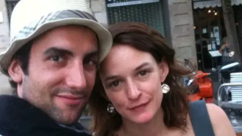 Courtesy of Audrey and Arthur Kermalvezen A close-up photograph shows a man and a woman - both with brown hair - posing together outdoors and looking toward the camera. The man wears a white hat. The backdrop is a city street with a building façade, outdoor seating and an orange scooter visible in the background.