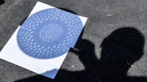Getty Images The silhouette of a man in shadow holding a colander over a piece of white paper as an alternative way to view a solar eclipse