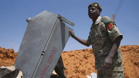 Los Angeles Times via Getty Images A man in uniform with a red beret stands in a hole filled with drone components  in El Obeid, Sudan - March 2026