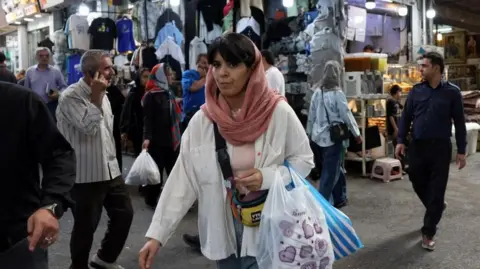 EPA A woman wearing a white shirt and pink scarf carries shopping bags on her left arm in the middle of a market, where clothes can be seen hanging and other shoppers walk about