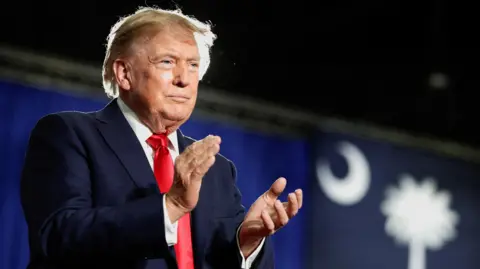 Reuters US President Donald Trump standing on a stage dressed in a navy suit and red tie clapping to an audience not in view. 
