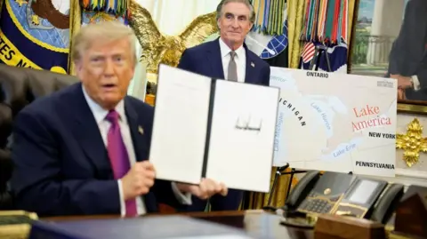 Reuters US President Donald Trump holds up a document with his signature in the Oval Office. Begin him is Secretary of the Interior Doug Burghum and a large map showing the US and Canada and the Great Lakes, with Lake Ontario renamed in red script as Lake America. 
