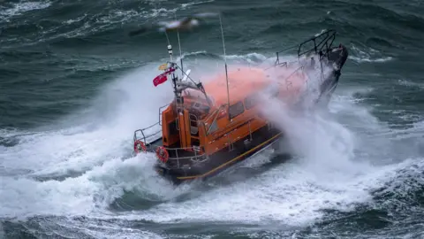 RNLI A Shannon class lifeboat in rough swell with a wave breaking over it.