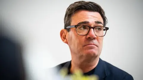 PA Media A headshot of Andy Burnham, wearing a black suit over a black t-shirt and black spectacles. He has a serious expression on his face.