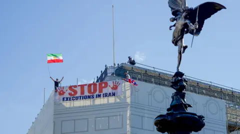 LightRocket via Getty Images Protesters hold a banner saying "stop executions in Iran" and wave an Iranian flag during a demonstration on the roof of the Trocadero, overlooking Piccadilly Circus, central London (30 July 2026)