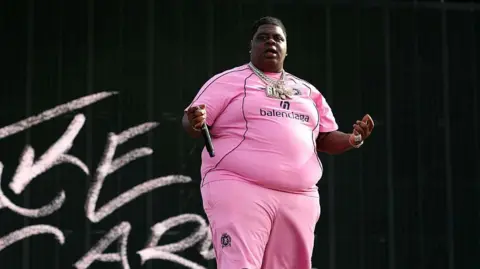 Getty Images The rapper wearing a pink outfit with 'balenciaga' written on the chest, and several necklaces, holds a microphone in his extended right hand away against a black background with some chalk-like letters to his right 