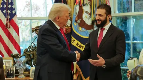 AP Two men in dark suits and red ties smile and shake hands in front of windows and American flags