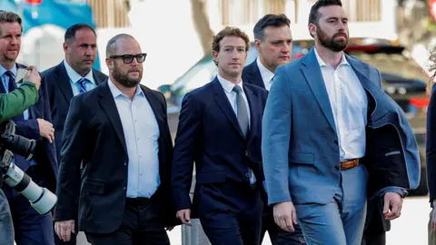 Reuters Mark Zuckerberg wearing a suit, looks towards the camera with a blank expression. He is flanked by staff also in suits. Photographed in February 2026