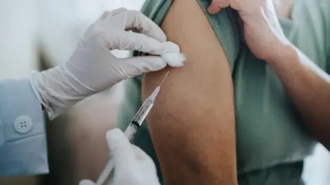 Getty Images Close-up of a healthcare professional administering an injection into a person's upper arm, with white gloved hands holding a syringe and cotton swab during a vaccination appointment.