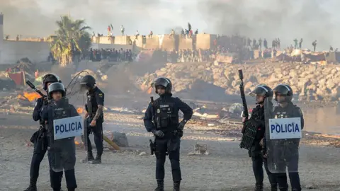 Getty Images Spanish armed police with riot shields on Trampolin beach, Ceuta, as anti-migrant protesters set fire to a migrant camp there. Their faces are blurred. Smoke can be seen rising in the air.