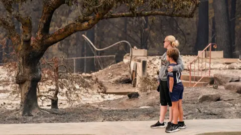 Reuters A woman dressed in black capris, grey jumper and black trousers wraps her arm around a young boy in black shorts, black top and black trainers, as both survey the damage of a burnt tree and rubble of a building