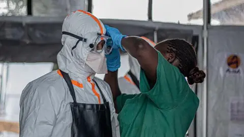 Reuters A health worker in green scrubs helps another person put on their PPE. The person on the left of the picture is wearing full white protective gear and goggles.