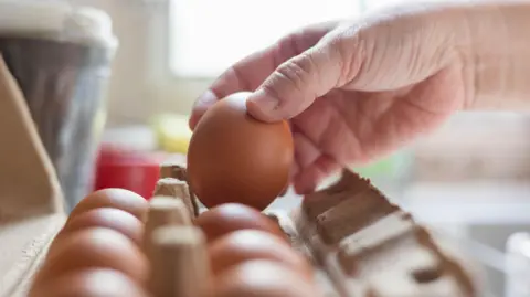 Getty Images A carton of eggs are in focus, with a hand picking up on egg.